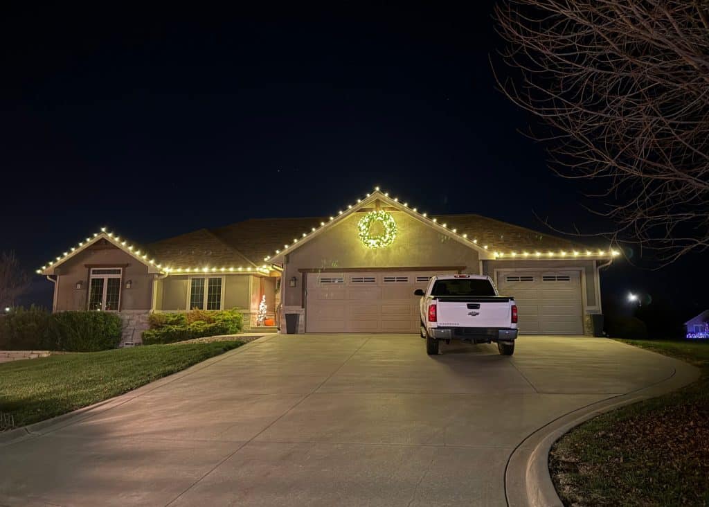 A house at night features holiday lighting with white lights along the roof and a large lit wreath above the garage. A white pickup truck is parked in the driveway, while the yard and leafless trees complete this festive, welcoming scene.