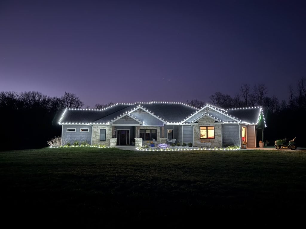 A single-story house is outlined with permanent lighting along the roof at dusk. The windows emit a warm glow, and a lawnmower is parked on the grass to the right. Trees are silhouetted against the dark sky in the background.