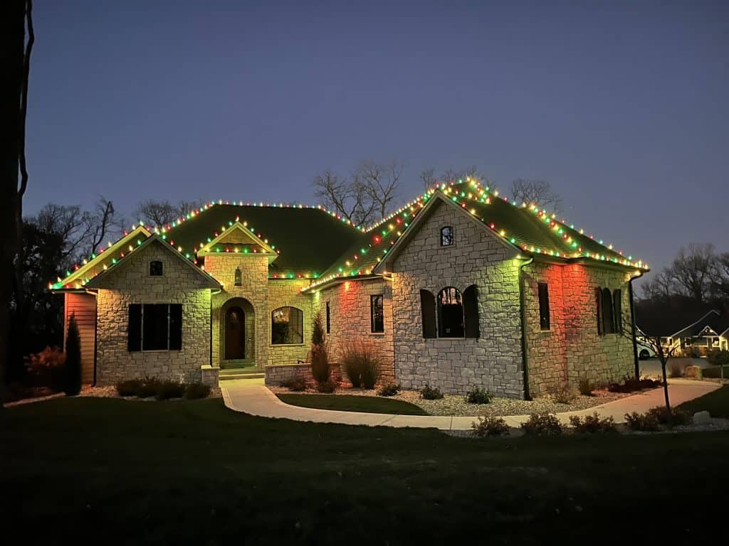 A stone house decorated with vibrant holiday lighting along the roofline is illuminated at dusk, with bare trees in the background and a winding walkway leading to the front door.