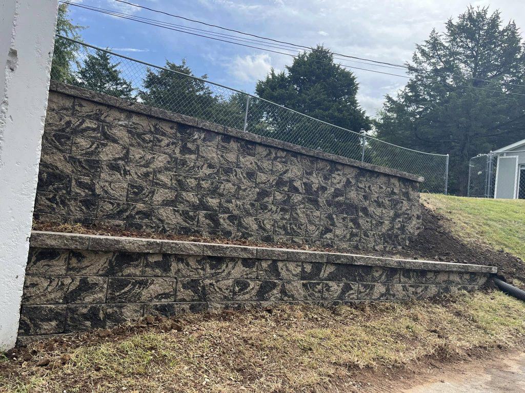 A landscape showcasing retaining walls with a patterned design features brown grass in front and a chain-link fence atop the structure. In the background, trees stand tall against a cloudy sky.