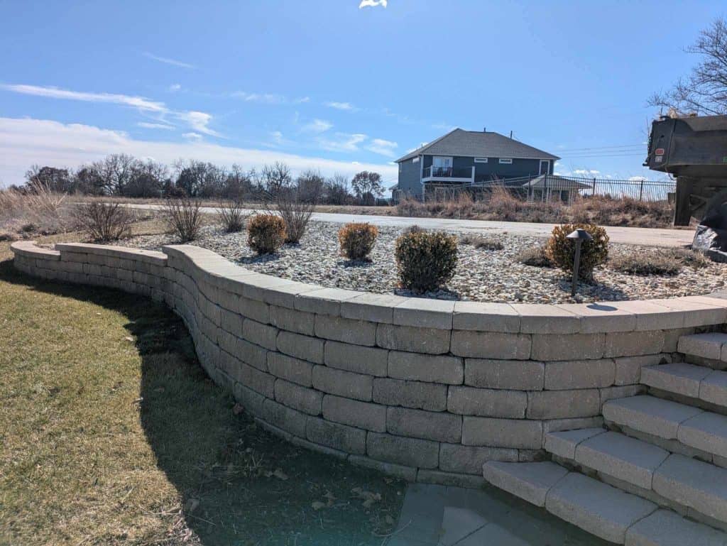 Stone retaining wall with a curved design, small shrubs planted in gravel atop the wall. Steps on the right lead upwards, showcasing excellent landscape maintenance. A house and a truck are visible in the background under a clear blue sky.