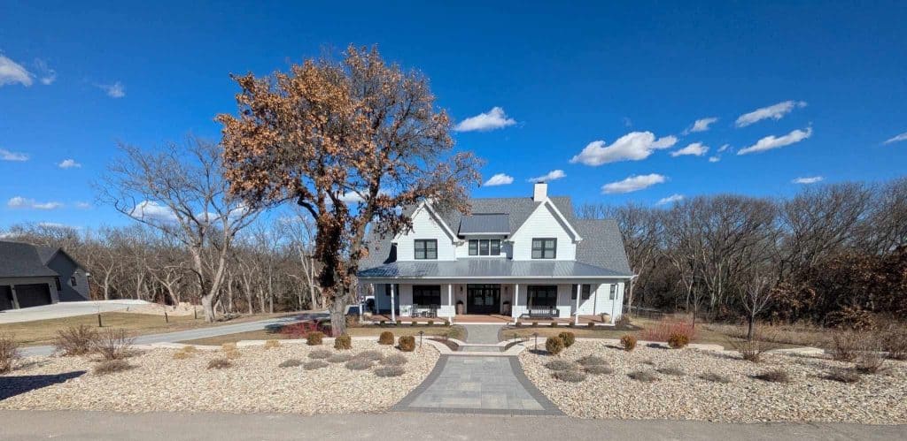 A house with a long driveway is surrounded by a stone garden and trees, showcasing meticulous landscape maintenance. A person stands in the foreground, casting a shadow while taking a photo. The sky is clear and blue with scattered clouds.