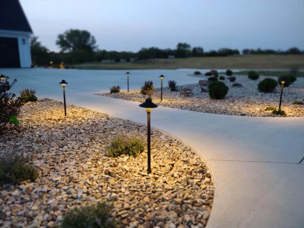 A gently curved concrete path winds through the outdoor landscape, adorned with small shrubs and decorative rocks. The path is illuminated by warm-lit pathway lights, enhancing the cozy ambiance at dusk.