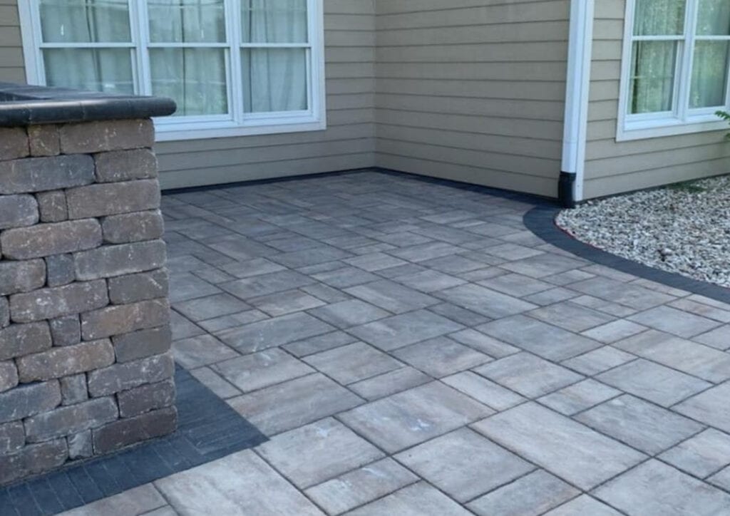 A backyard patio with gray rectangular paver patios stones, bordered by dark bricks. A stone wall is on the left, and beige siding with white-trimmed windows and a section of white gravel is on the right.