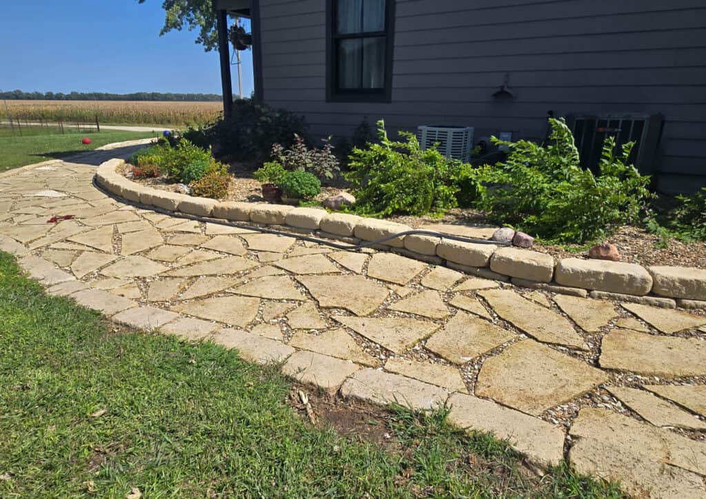 A stone walkway curves beside a house, bordered by a raised stone flower bed filled with green plants and flowers. Grass grows next to the path, with paver patios seamlessly blending into the landscape under a clear blue sky.