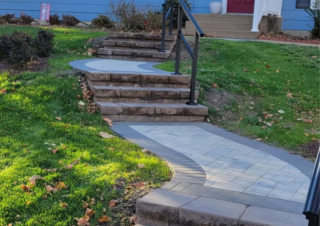 Curved stone walkway with patterned pavers blends into a small flight of stone steps and black railing, bordered by green grass and shrubs, leading to a blue house—ideal inspiration for elegant paver patios.
