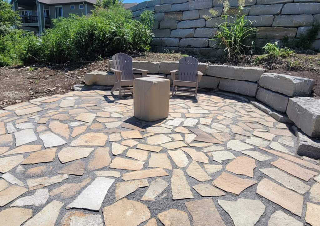 Two wooden chairs face a square stone fire pit on a circular paver patio surrounded by low stone walls and lush green plants, with houses and a hill visible in the background.