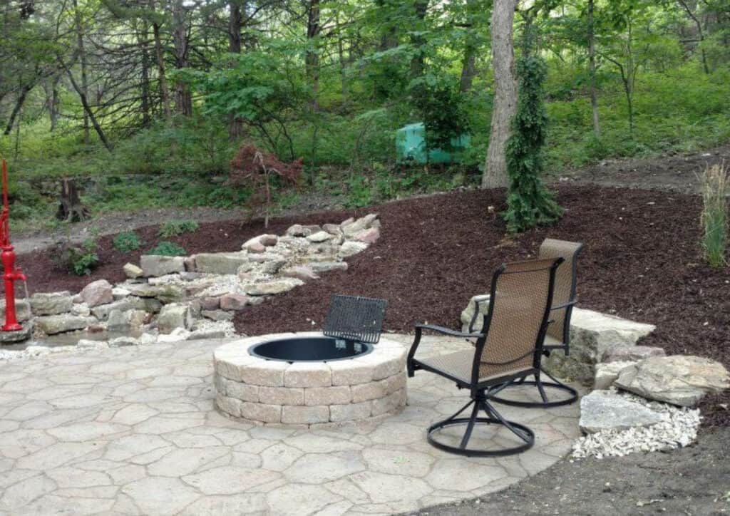 A stone paver patio with two metal chairs faces a circular stone fire pit. Behind the patio, rocks and mulch form landscaping, surrounded by trees and greenery in a wooded area.