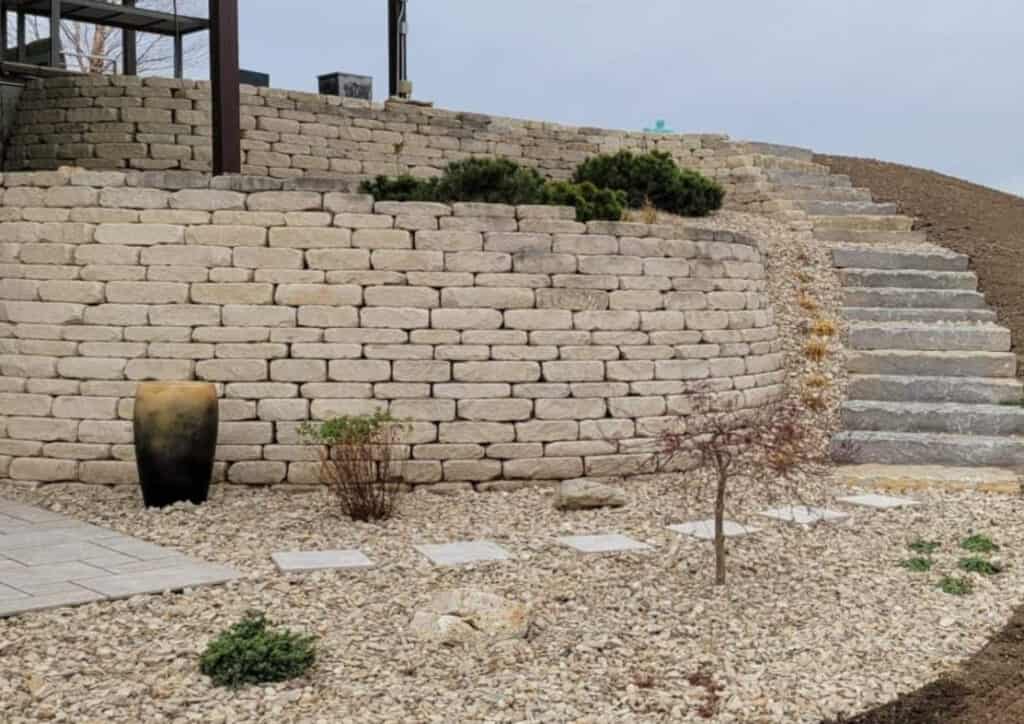 A terraced stone retaining wall with steps leads up a slope. The landscaping features gravel, pavers, small shrubs, a decorative pot, and sparse plants. Overcast skies set the backdrop for these functional and stylish retaining walls.