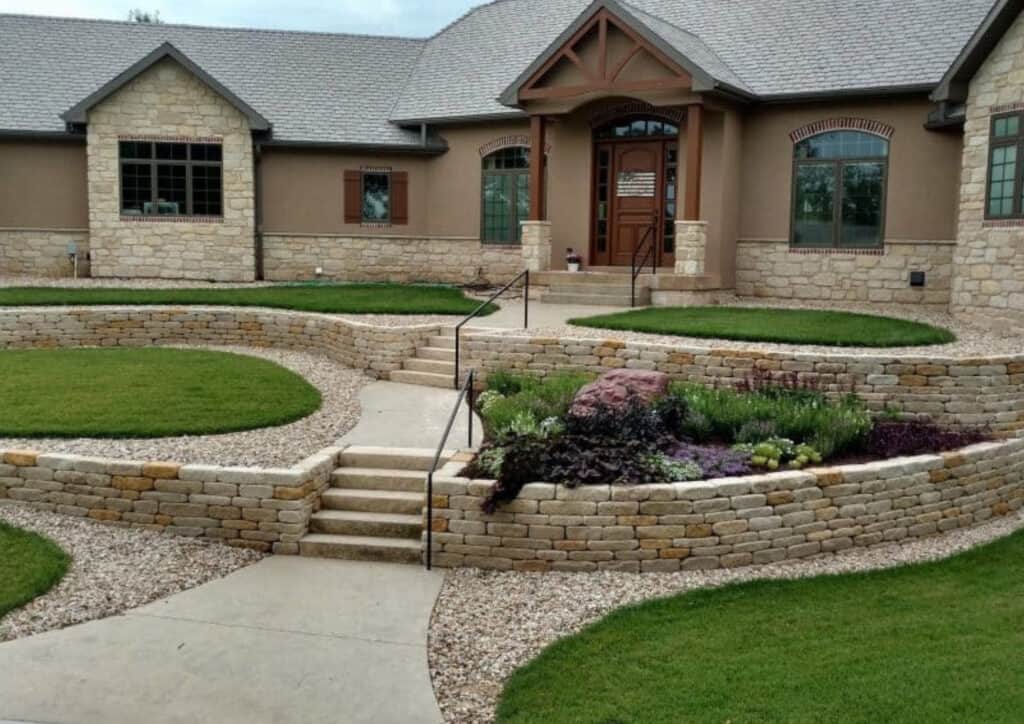 A modern house with tan stone and stucco exterior features elegant retaining walls, a landscaped yard, curved stone details, steps with a handrail, and green lawns with a central flower bed in front.