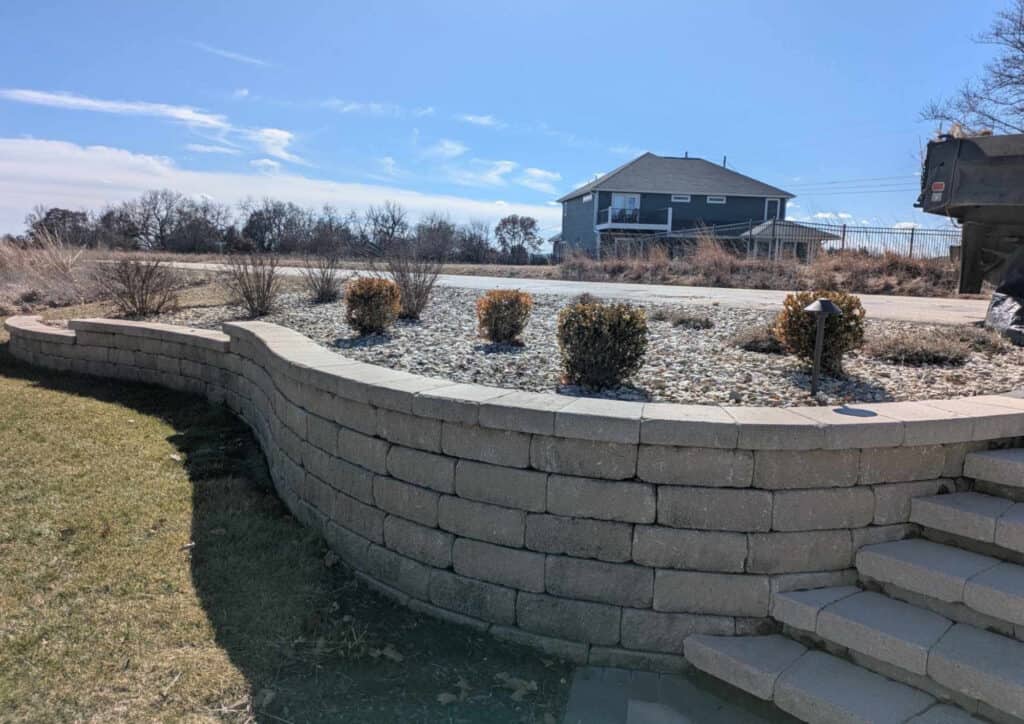 A curved stone retaining wall with steps leads to a gravel-covered area dotted with small shrubs. Retaining walls frame the scene, with a grassy lawn in the foreground and a blue house set against a clear, sunny sky.