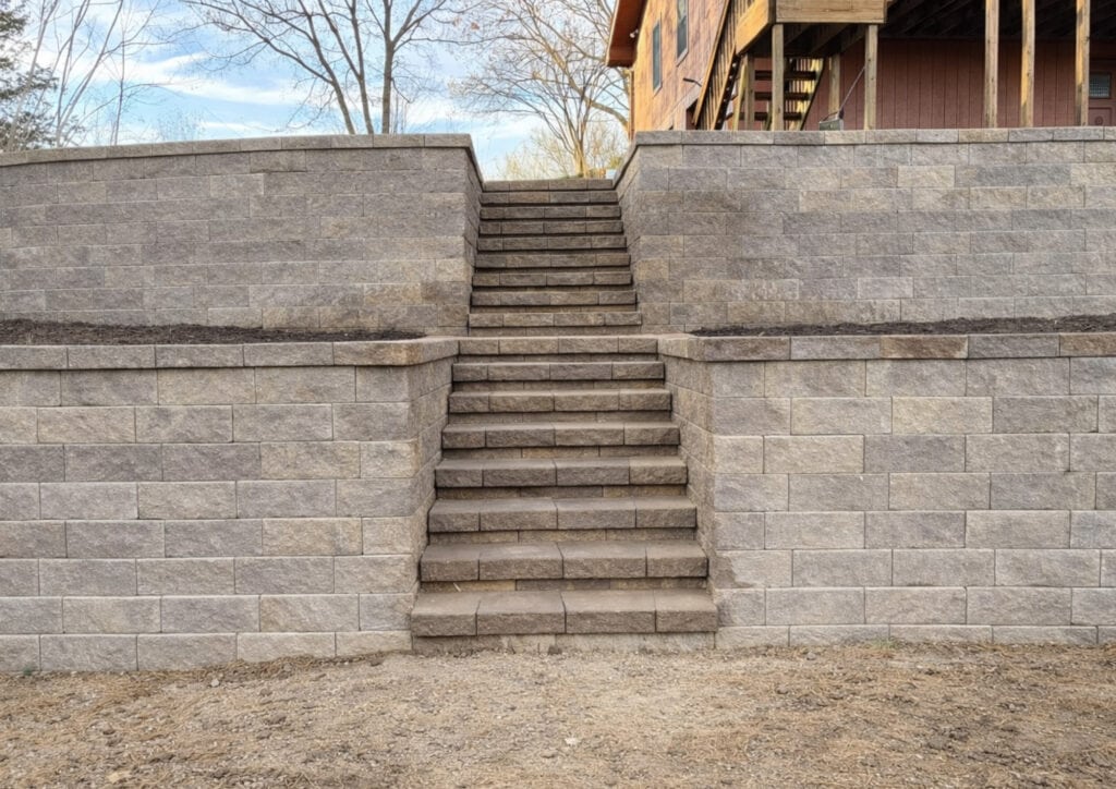 Stone steps lead up between two gray retaining walls, with soil beds on top. The stairs ascend to a partially visible wood structure—possibly a deck or house—with bare trees silhouetted in the background.