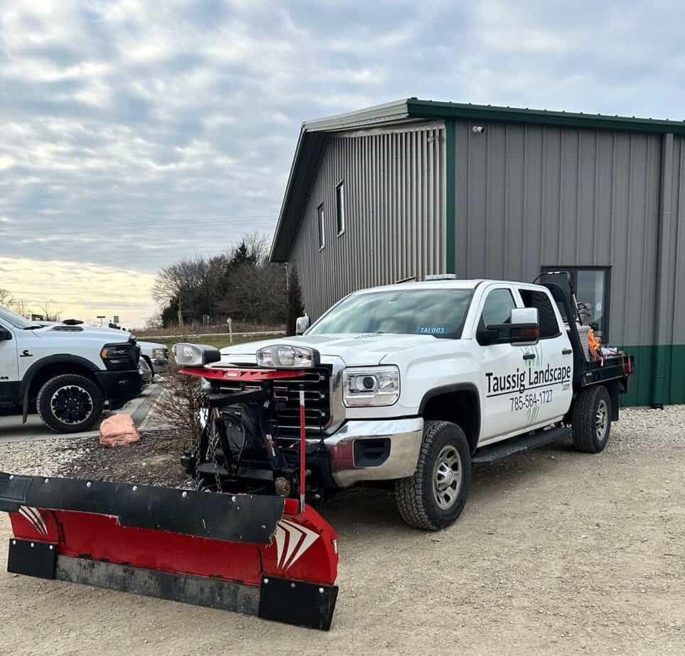 A white pickup truck with a snowplow attachment, part of Taussig Landscape's Snow and Ice Removal fleet, is parked on a gravel surface in front of a metal building. Another vehicle is partially visible to the left under the cloudy sky.