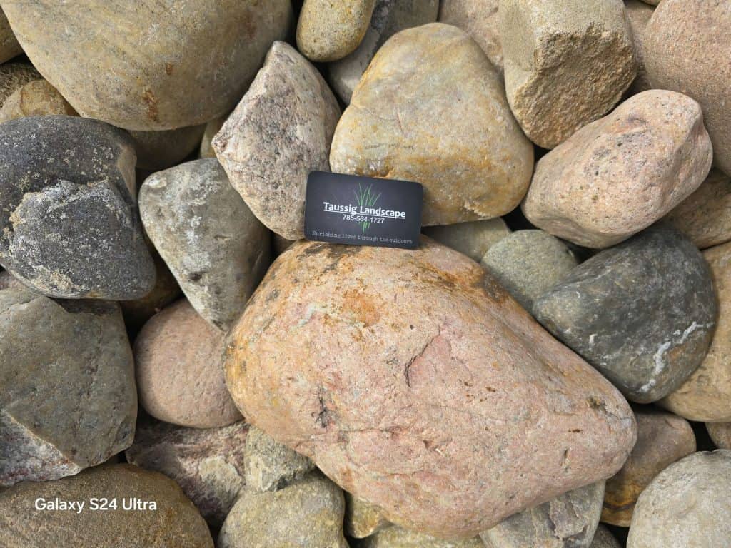 A business card from "Taussig Landscape" is placed among variously sized and colored cobble stones. The card has contact information and the slogan, "Breathing New Life Through Your Landscape." The words "Galaxy S24 Ultra" are visible in the bottom left corner of the image.