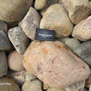 A business card from "Taussig Landscape" is placed among variously sized and colored cobble stones. The card has contact information and the slogan, "Breathing New Life Through Your Landscape." The words "Galaxy S24 Ultra" are visible in the bottom left corner of the image.