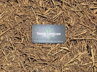 A business card for "Taussig Landscape" lies on a bed of red mulch. The card includes a phone number (785-564-1772) and the tagline "Enriching lives through the outdoors." The card features the company name and a green logo representing grass, reminiscent of scenic Ozark landscapes.