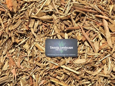 A business card for Taussig Landscape, featuring their phone number and tagline, is elegantly placed on a bed of cedar mulch. The card, with its black background and white text complemented by a graphic of green grass, highlights the natural mulch benefits they offer.