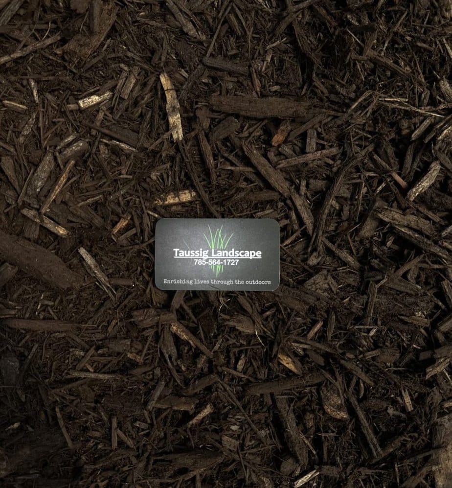 A small business card reading "Taussig Landscape Corp." is placed on rich brown colored mulch, surrounded by pieces of wood and organic material.