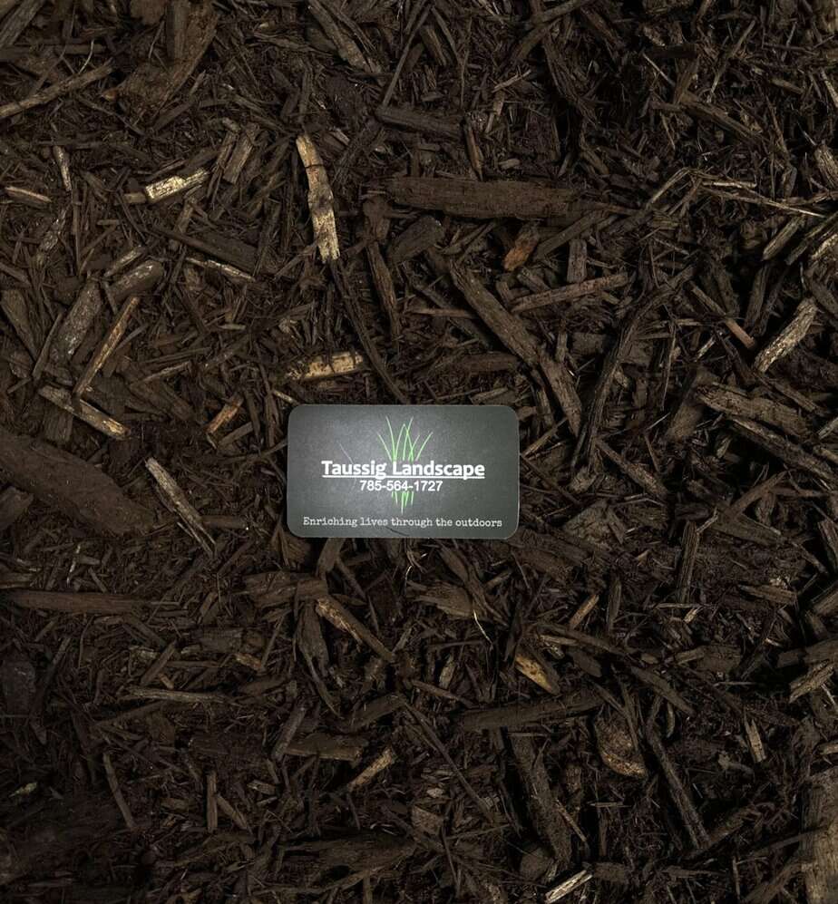 A small business card reading "Taussig Landscape Corp." is placed on rich brown colored mulch, surrounded by pieces of wood and organic material.