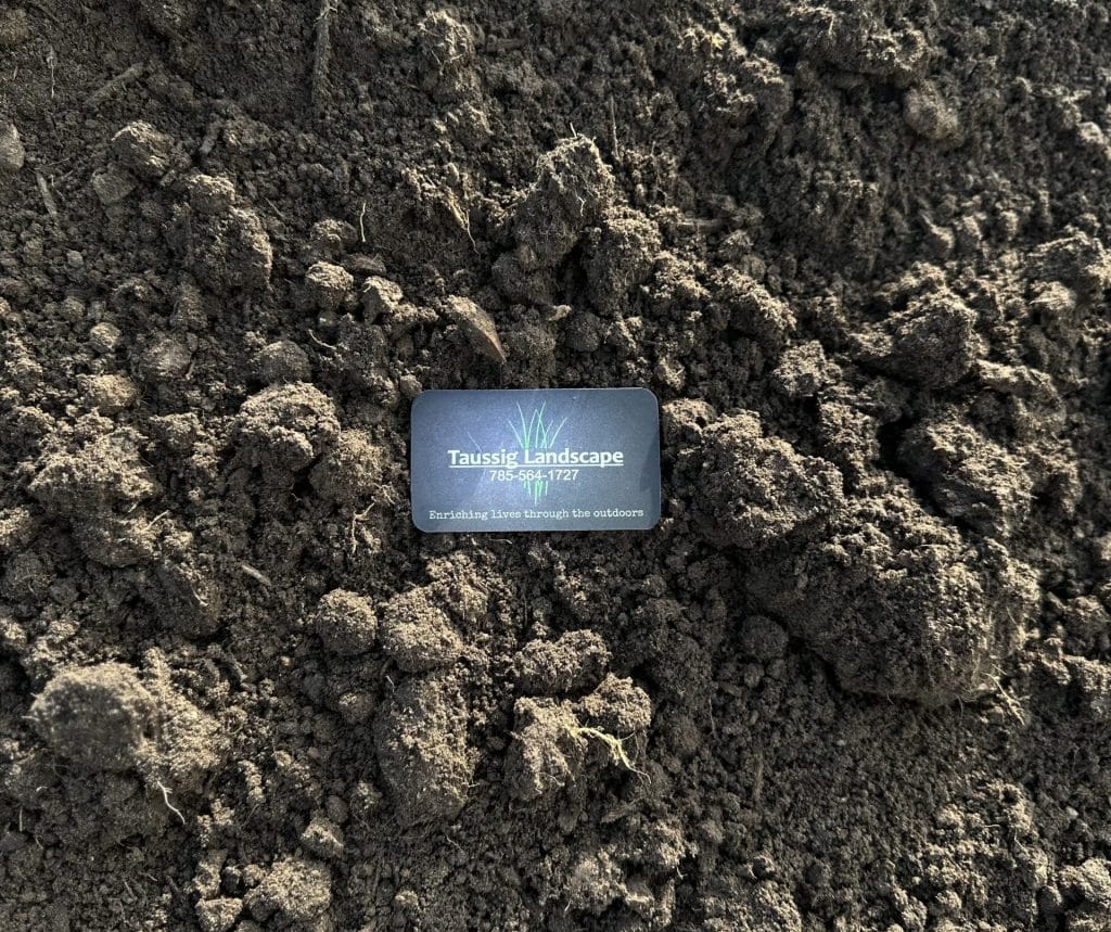 A business card for Tussis Landscape Ltd sits on uneven, dark brown soil with clumps and small ridges, highlighting the earthy texture and rough surface of the ground.