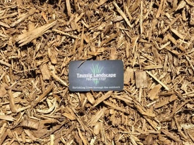 A business card for Taussig Landscape, displaying a phone number, is placed on a heap of brown wood mulch. The card features green grass graphics and reads, "Enriching lives through the outdoors.
