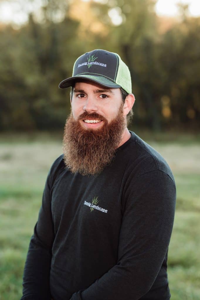 A bearded man wearing a black long-sleeve shirt and a cap with a logo, standing outdoors in a grassy area with trees in the background.