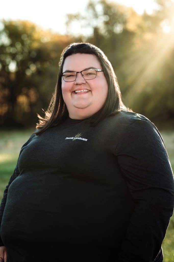 A person with long hair and glasses smiles while standing outdoors. They are wearing a dark long-sleeve shirt, and sunlight filters through the trees in the background.