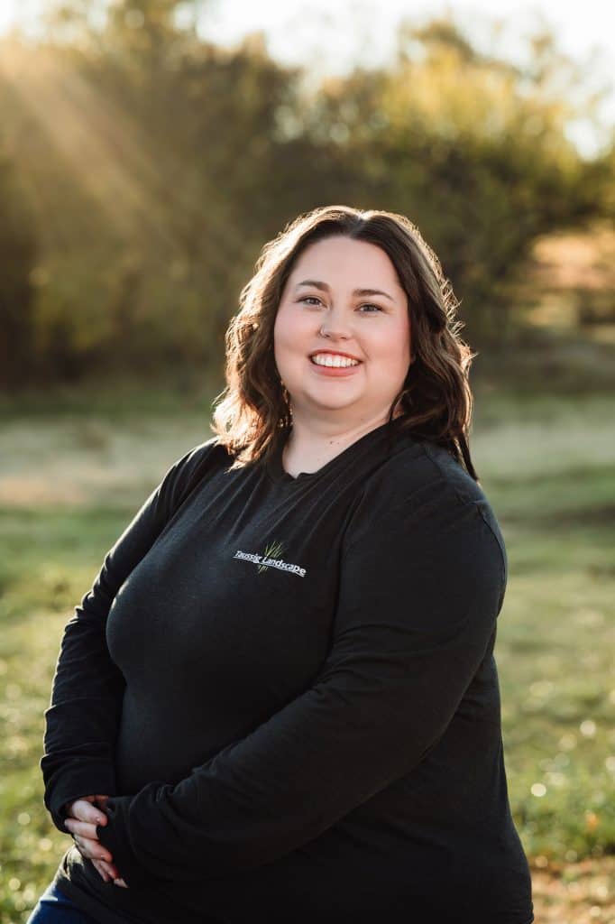 A woman with long brown hair and a black long-sleeve shirt smiles while seated outdoors. Sunlight filters through trees in the background, creating a warm, natural setting.