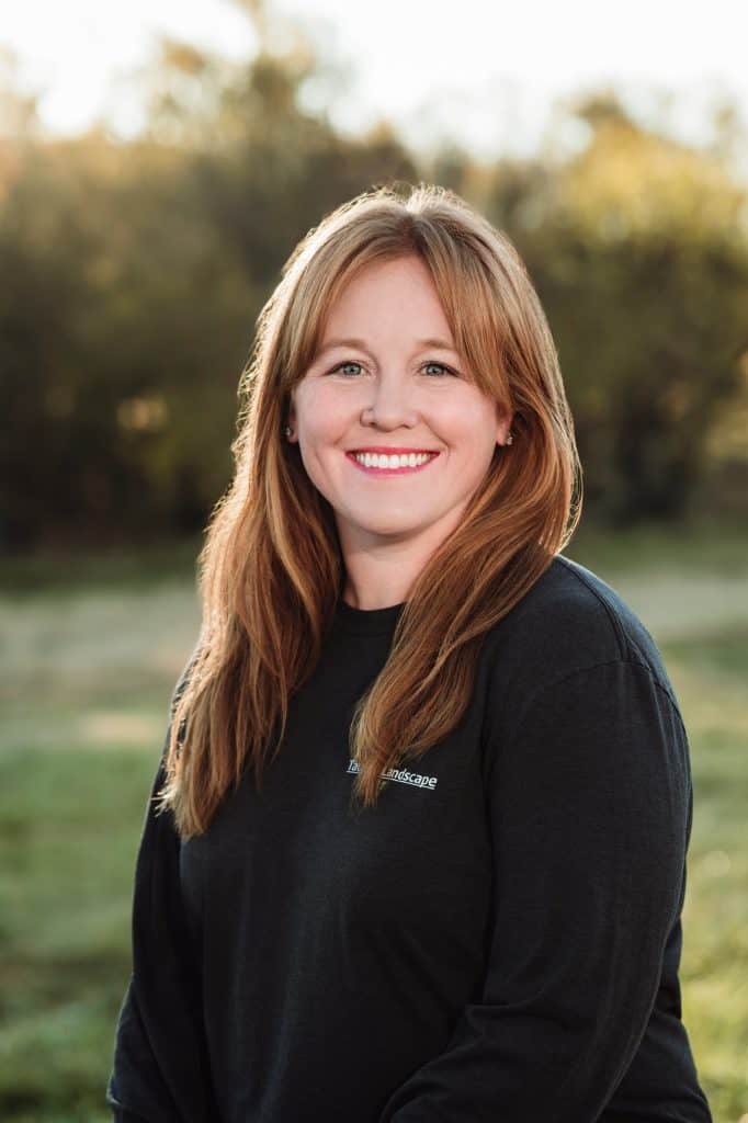 A woman with long, reddish-brown hair smiles while standing outdoors in a natural setting with trees and grass in the background. She is wearing a black long-sleeved shirt. The sunlight provides a soft, warm glow.