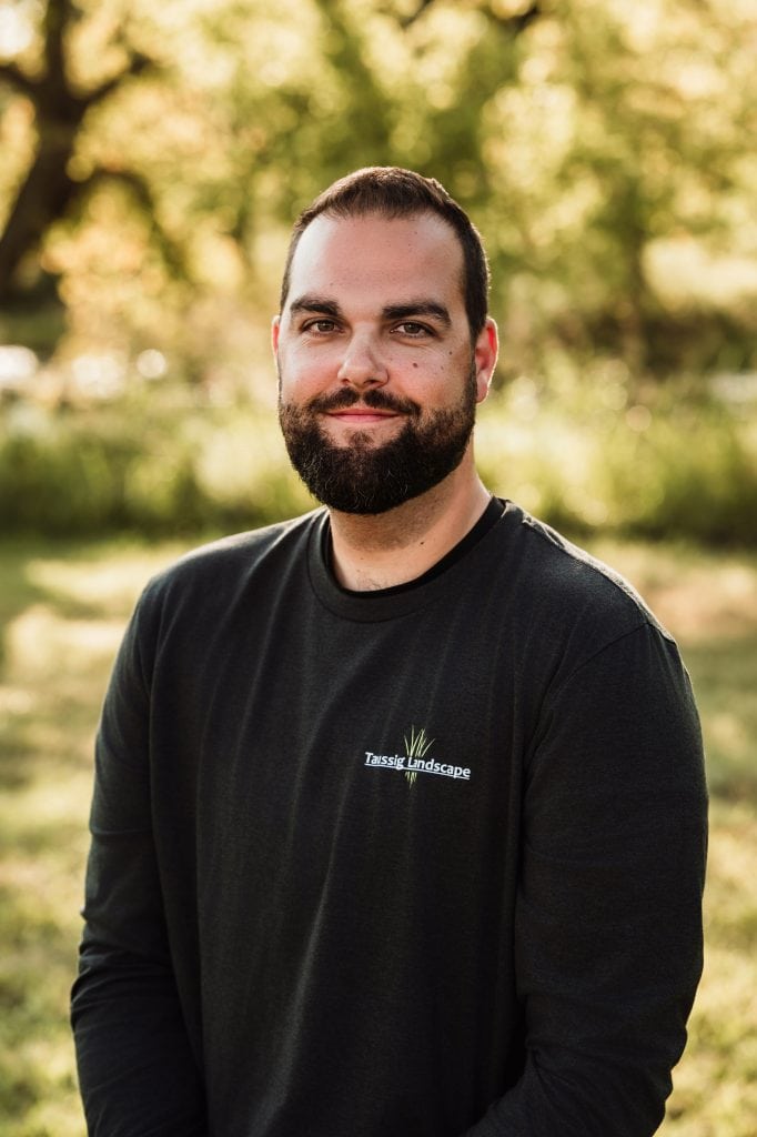 Kevin Albrecht, a man with a beard and short dark hair, stands outside in a sunlit, grassy area wearing a black long-sleeve shirt with the "Tessa Landscape" logo. Trees create a natural backdrop behind him.