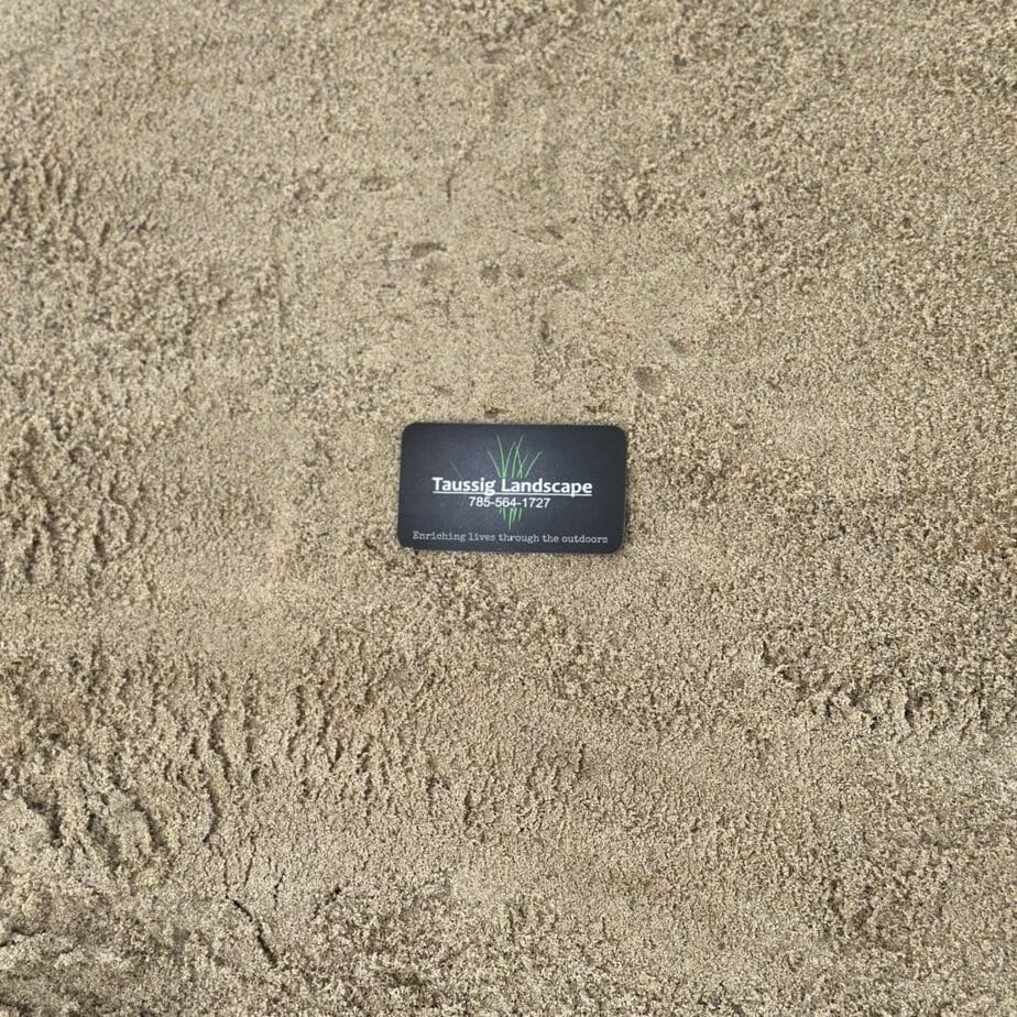 A black business card with the text "Talasek Landscape 512-944-7434, Everlasting Outdoor Living" lies on a sandy surface. The card features a small graphic of grass.