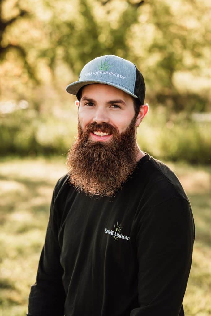 A bearded man, Josh Schraad, wearing a blue and black cap and a black shirt with a "Texas Landscape" logo, stands outdoors in a sunlit, grassy area with trees in the background, smiling at the camera.