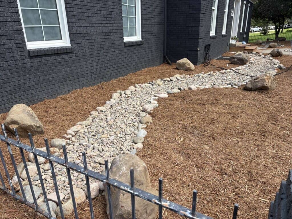 A landscaped garden bed with a dry creek made of light-colored stones for erosion control, bordered by large rocks, sits beside a dark-brick house with white-trimmed windows and brown mulch covering the ground.