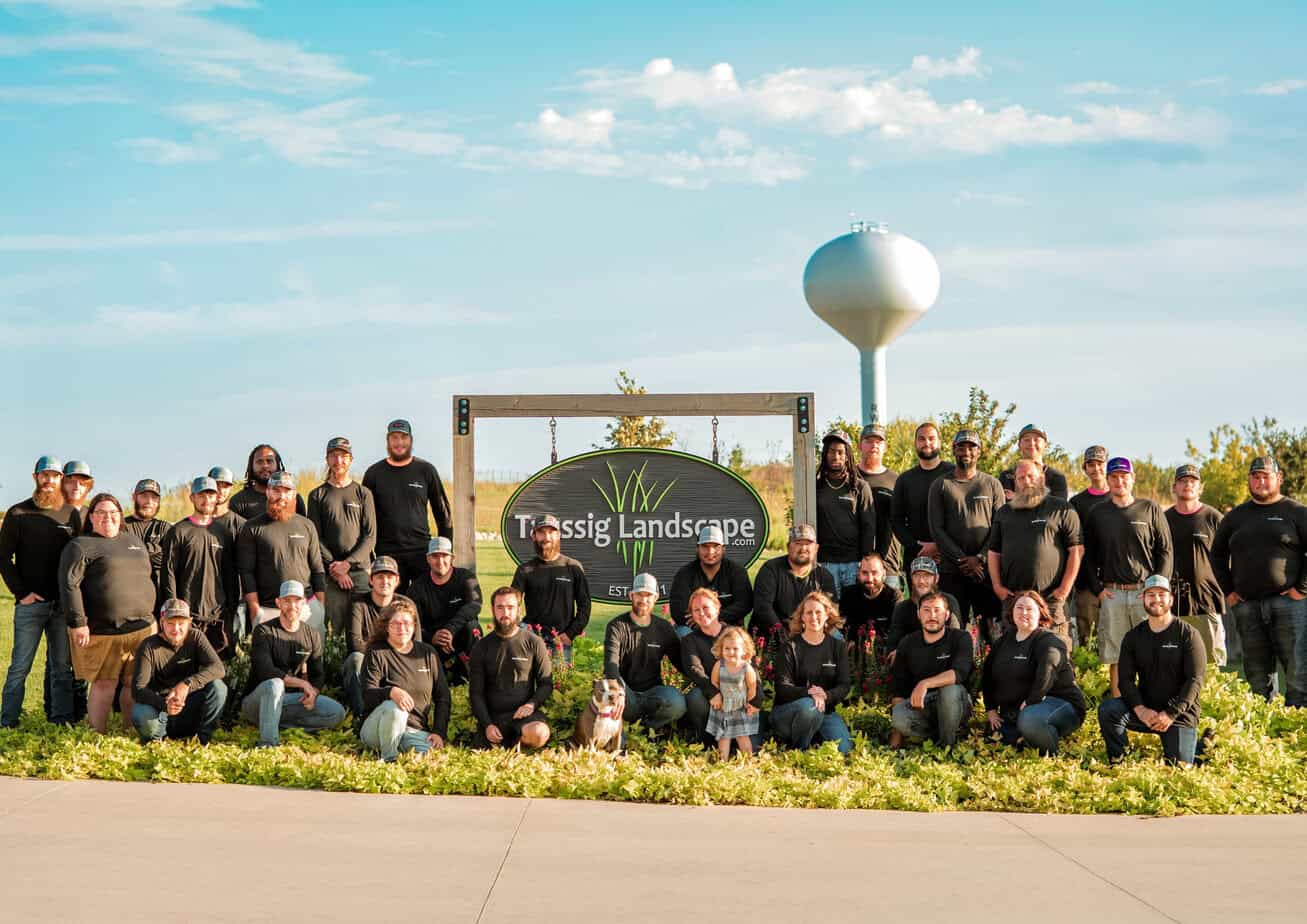 A large group of people in matching black shirts pose together outdoors in front of a "Tessig Landscape" sign, with greenery, a water tower, and a blue sky in the background.