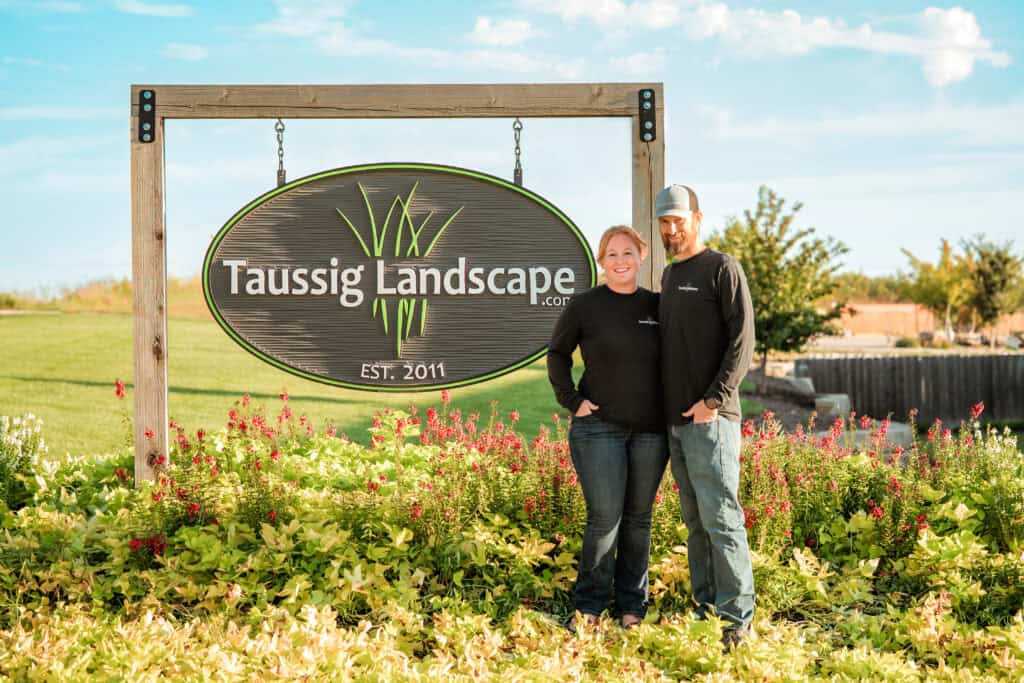 Two people stand smiling in front of a large "Taussig Landscape" sign surrounded by green grass and colorful plants, with a blue sky in the background.