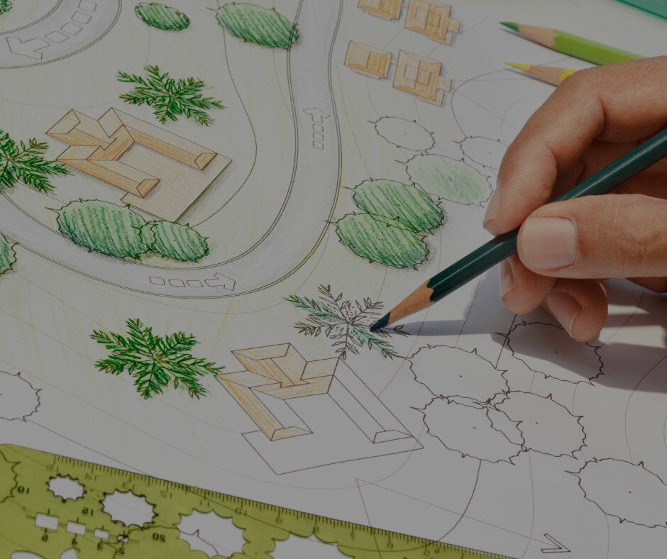 A hand uses a green colored pencil to draw trees on an architectural site plan, demonstrating the role of a Landscape Design Assistant amidst drawing tools and other colored pencils, enhancing buildings, paths, and landscaping details.